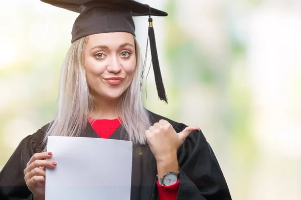 指していると幸せそうな顔を笑顔で側まで親指で示す分離の背景上の学位を保持する大学院の制服を着て若いブロンドの女性 — ストック写真