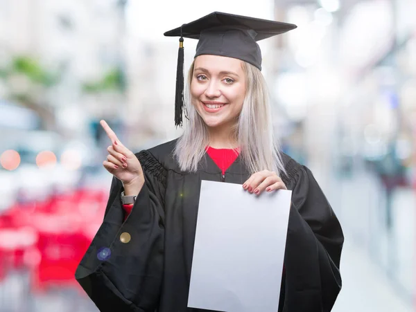 若いブロンドの女性側の手と指で指す非常に幸せな孤立の背景の上の学位を保持する大学院の制服を着て — ストック写真