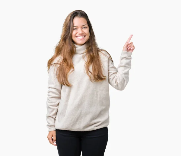 若い美しいブルネットの女性の顔に大きな笑みを浮かべて隔離された背景にタートルネックのセーターを着てカメラを見て側の手と指で指す — ストック写真