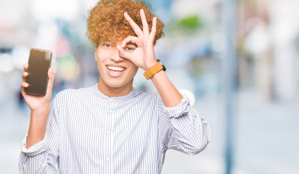 指を通して見る目に手で サインをして笑っている幸せそうな顔で若いハンサムな男を示すスマート フォンの画面 — ストック写真