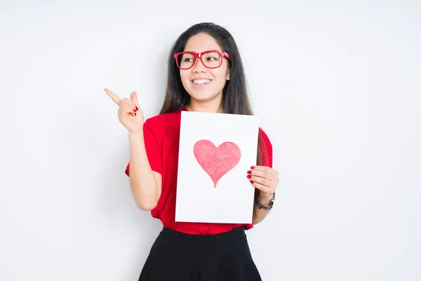 Young brunette woman holding card with red heart over isolated background very happy pointing with hand and finger to the side