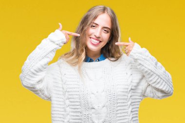 Kendinden emin gösterilen ve işaret parmakları diş ve ağız ile gülümseyen izole arka plan üzerinde kış kazak giyen güzel genç sarışın kadın. Sağlık kavramı.