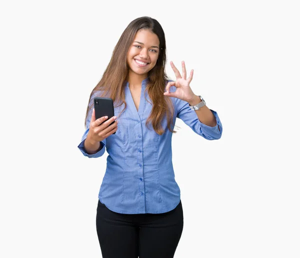 年轻美丽的黑发女商人使用智能手机在孤立的背景做确定的标志与手指 优秀的象征 — 图库照片