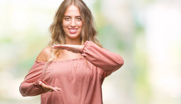 Beautiful young blonde woman over isolated background gesturing with hands showing big and large size sign, measure symbol. Smiling looking at the camera. Measuring concept.