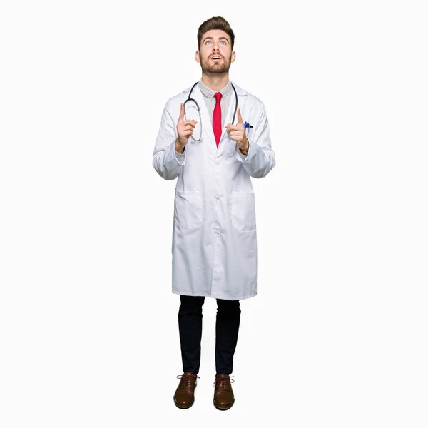 医療のコートを着ている若いハンサムな医者男びっくりし 驚き見て 指で指して 腕を上げた — ストック写真