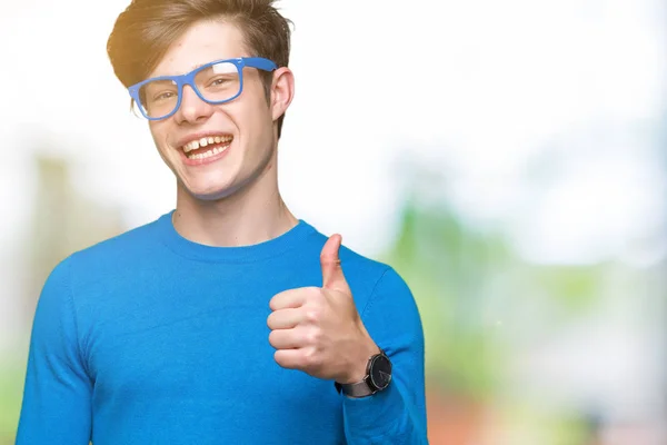 年轻英俊的男人戴着蓝色的眼镜在孤立的背景做快乐的大拇指用手的手势 通过显示成功来批准镜头的表达 — 图库照片