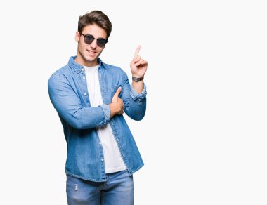 Kameraya bakarak yan el ve parmak ile işaret eden genç yakışıklı adam yüzünde kocaman bir gülümseme ile izole arka plan üzerinde güneş gözlüğü takıyor.