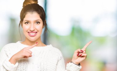 Kış süveteri giyen, gülümseyen ve kameraya bakan genç ve güzel bir kadın iki eliyle ve parmaklarıyla kenarı işaret ediyor..