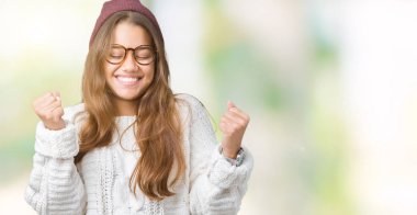 Genç güzel esmer hipster kadın gözlük ve kış şapka giymek izole arka plan üzerinde başarı için heyecanlı silah ile zafer gülümseyen kutlamak kaldırdı. Kazanan kavramı.