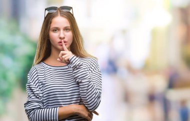 Genç güzel sarışın kadın dudaklarında parmak ile sessiz olmanı izole arka plan üzerinde güneş gözlüğü takıyor. Sessizlik ve gizli kavramı.