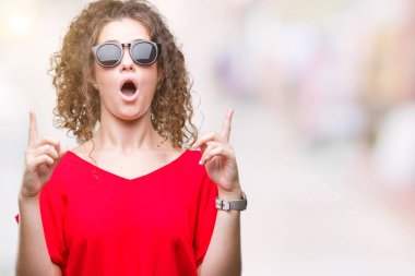 Güzel esmer kıvırcık saçlı Genç kız izole arka plan üzerinde güneş gözlüğü takıyor hayret ve yukarıya bakıp parmakları ile işaret şaşırttı ve silah kaldırdı.