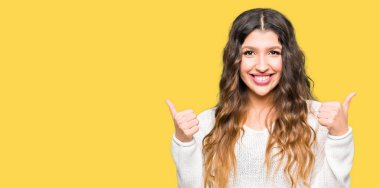 Beyaz kazak başarı işareti el ile pozitif jest yapıyor giyen genç güzel kadın başparmak gülümseyen ve mutlu. Neşeli bir ifadeyle, kazanan jest kameraya bakıyor.