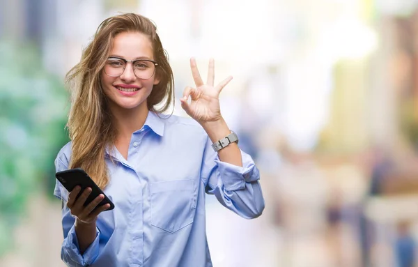 年轻美丽的金发女商人使用智能手机在孤立的背景做确定的标志与手指 优秀的象征 — 图库照片