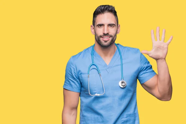 英俊的年轻的医生外科医生在孤立的背景显示和指向用手指数字五 同时微笑自信和快乐 — 图库照片
