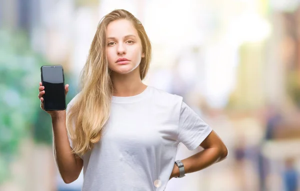 年轻美丽的金发女子显示屏幕的智能手机在孤立的背景与自信的表情在聪明的脸思考严重 — 图库照片