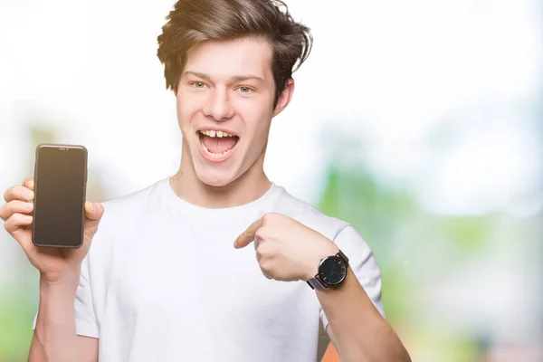 Young man showing smartphone screen over isolated background with surprise face pointing finger to himself