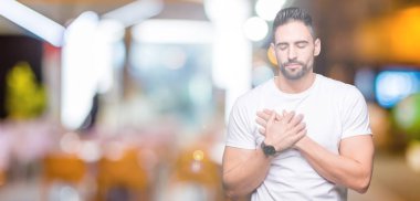 Genç adam ile gülümseyen izole arka plan üzerinde rahat beyaz t-shirt giyen kapalı gözler ve yüzünde minnettar jest ile göğüs üzerinde eller. Sağlık kavramı.