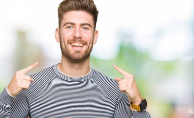 Kendinden emin gösterilen ve işaret parmakları diş ve ağız ile gülümseyen çizgili süveter giymiş yakışıklı delikanlı. Sağlık kavramı.