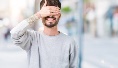 Genç yakışıklı adam gülümsüyor ve gözleri için sürpriz kapsayan yüz el ile gülüyor izole arka plan üzerinde kazak giyiyor. Kör kavramı.