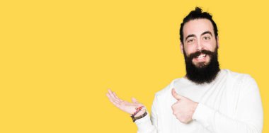 Mutlu ve neşeli gülümseyen genç hipster adamla uzun saç ve sakal sportif kazak gösteren palm el giyen ve, baş parmakları Tamam jest yapıyor