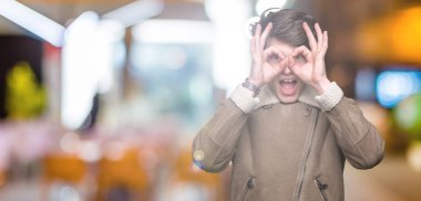 Yakışıklı delikanlı üzerinde kış palto giyiyor, dilini parmaklarının arasından baktığını yapışmasını dürbün gibi ok jest yapıyor arka plan izole. Deli ifade.