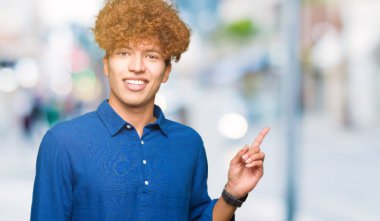 Yüzünde kocaman bir gülümseme ile afro saçlı genç yakışıklı zarif adam kameraya bakarak yan el ve parmak ile işaret.