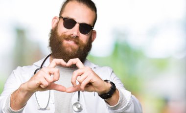Kalp simgesi ve elleriyle şekil gösterilen aşık gülümseyen güneş gözlüğü takıyor sakallı genç sarışın doktor adam. Romantik kavramı.
