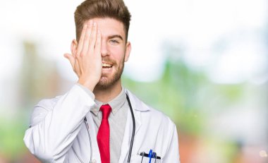 Tıbbi giyen genç yakışıklı Doktor adam kat yüz ve sürpriz duygu kendinden emin gülümseme ile el ile bir göz kapsayan.
