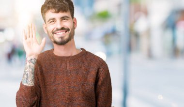 Yakışıklı delikanlı izole arka plan Waiving mutlu selam söyleyerek üzerinde kış kazak ve gülümseyen, dostça karşılama jest