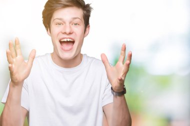 Üzerinde rahat beyaz t-shirt giyen genç yakışıklı adam deli kutluyor arka plan izole ve başarı için kaldırdı kollarını açarak hayrete ve heyecanlı çığlık gözleri açık. Kazanan kavramı