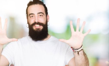 Uzun saç ve sakal gösterilen ve parmakları ile işaret casual beyaz t-shirt giyen genç hipster adamla kendinden emin ve mutlu gülümseyerek süre on numara.