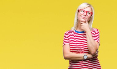 Genç güzel sarışın kadın gözlük ile izole arka plan üzerinde çene düşünme hakkında soru, dalgın ifade ver. Düşünceli yüzüyle gülümsüyor. Şüphe kavramı.