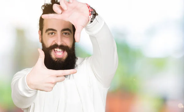 Young man with long hair and beard wearing sporty sweatshirt smiling making frame with hands and fingers with happy face. Creativity and photography concept.