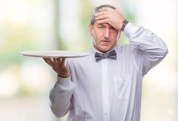 Handsome senior waiter man holding silver tray over isolated background stressed with hand on head, shocked with shame and surprise face, angry and frustrated. Fear and upset for mistake.