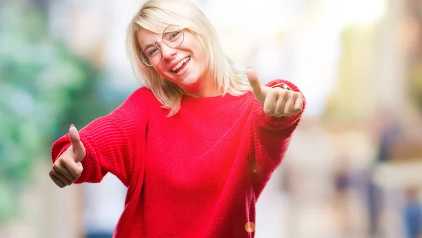 Young beautiful blonde woman wearing sweater and glasses over isolated background approving doing positive gesture with hand, thumbs up smiling and happy for success. Looking at the camera, winner gesture.