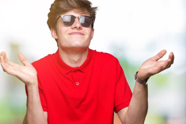 Genç yakışıklı adam izole arka plan clueless ve şaşkın bir ifadeyle kollar ve eller kaldırdı üzerinde güneş gözlüğü takıyor. Şüphe kavramı.