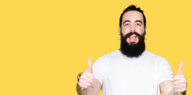 Uzun saç ve sakal el ile olumlu hareketi yaparken rahat beyaz t-shirt başarı işareti giyen genç hipster adamla başparmak gülümseyen ve mutlu. Neşeli bir ifadeyle, kazanan jest kameraya bakıyor.