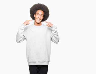 Afrikalı-Amerikalı genç afro saçlı emin gösterilen ve işaret parmakları diş ve ağız ile gülümseyen sportif kazak giyiyor. Sağlık kavramı.