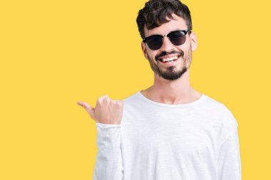 Yakışıklı delikanlı arıyor ve başparmak yanında yukarıyı mutlu yüz ile gülümseyen izole arka plan üzerinde güneş gözlüğü takıyor.