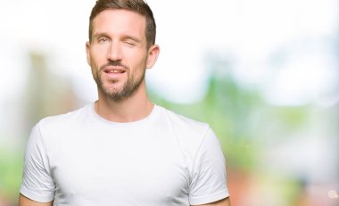 Casual beyaz t-shirt seksi bir ifadeyle, neşeli ve mutlu yüzünü kameraya bakarak göz kırpıyor giyen yakışıklı adam.