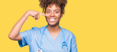 Genç Afro-Amerikan doktor kadın izole arka plan işaretçi el parmak yüz ve burun, neşeli gülümseyen