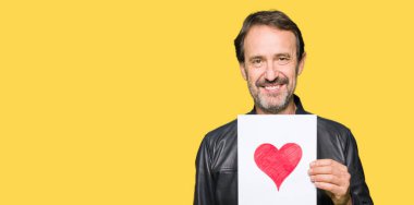 Ayakta ve diş gösterilen kendine güvenen bir gülümsemeyle gülümseyerek mutlu bir yüz ile kırmızı kalp ile kağıt tutan yakışıklı orta yaş erkek