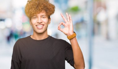 Afro saç giyen siyah t-shirt el ve parmaklar ok işaretiyle yapıyor olumlu gülümseyen yakışıklı delikanlı. Başarılı ifade.