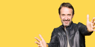 Orta yaş yakışıklı adam sarılmak için kollarını açarak gülümseyerek kameraya bakarak siyah deri ceket giyiyor. Neşeli ifade kucaklayan mutluluk.