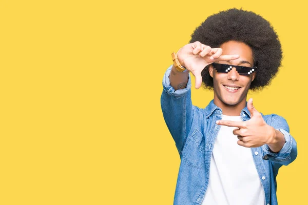 Young african american man with afro hair wearing thug life glasses smiling making frame with hands and fingers with happy face. Creativity and photography concept.