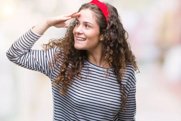 Beautiful brunette curly hair young girl wearing stripes sweater over isolated background very happy and smiling looking far away with hand over head. Searching concept.