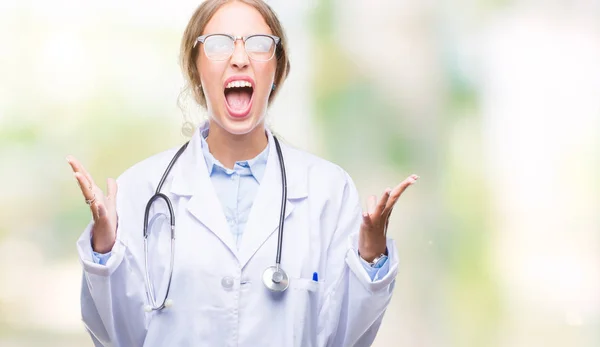 美丽的年轻金发碧眼的医生妇女穿着医疗制服在孤立的背景疯狂和疯狂的喊声和大喊大叫与侵略性的表情和手臂提出 挫折的概念 — 图库照片