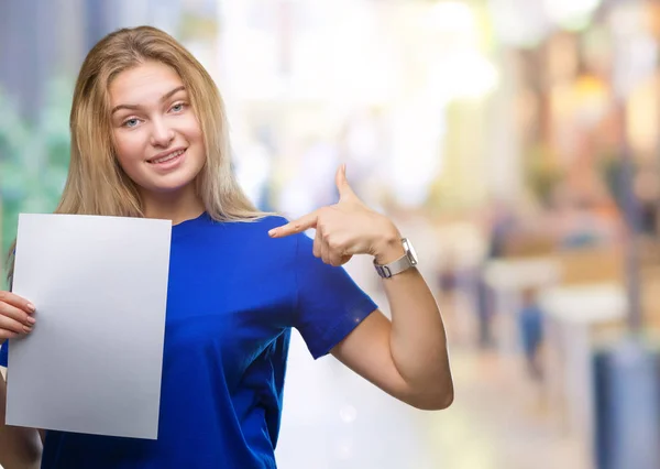 Young caucasian woman holding blank paper sheet over isolated background with surprise face pointing finger to himself