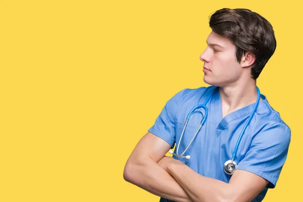年轻的医生穿着医疗制服在孤立的背景微笑着看着一边与胳膊横渡说服和自信 — 图库照片