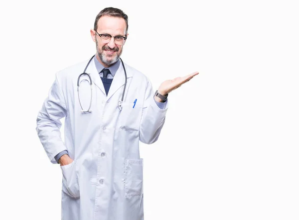 陽気な提示とカメラを見て手の手のひらで指している笑みを浮かべて医療制服孤立した背景を着て中年先輩の白髪医師男性 — ストック写真